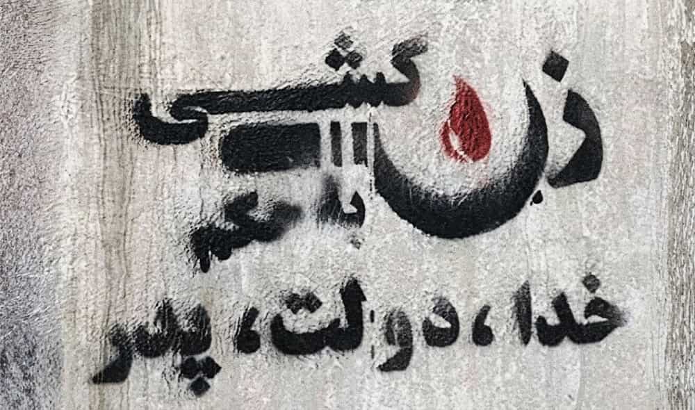 تصویری از یک دیوار خاکستری رنگ که روی آن شعاری به زبان فارسی با رنگ سیاه و قطره‌ای خون به رنگ قرمز استنسل شده است. متن نوشته شده: «زن کشی، به حکم خدا، دولت، پدر».